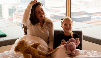 Mor og datter smiler og griner med en bog og bamser foran vindue med udsigt over byen