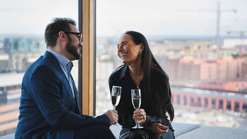 Mand og kvinde nyder hinandens selskab med champagne i hånden foran vindue med udsigt i hotellejlighed